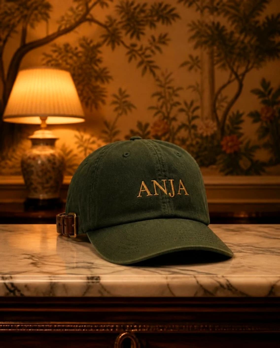 Washed green cap with ANJA embroidered in gold, on a marble console under a lamp