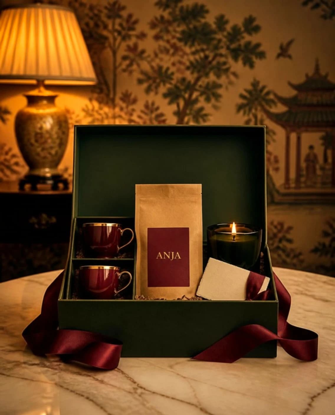 Green gift box with an oxblood ribbon, holding two espresso cups, a bag of coffee, a candle and a card