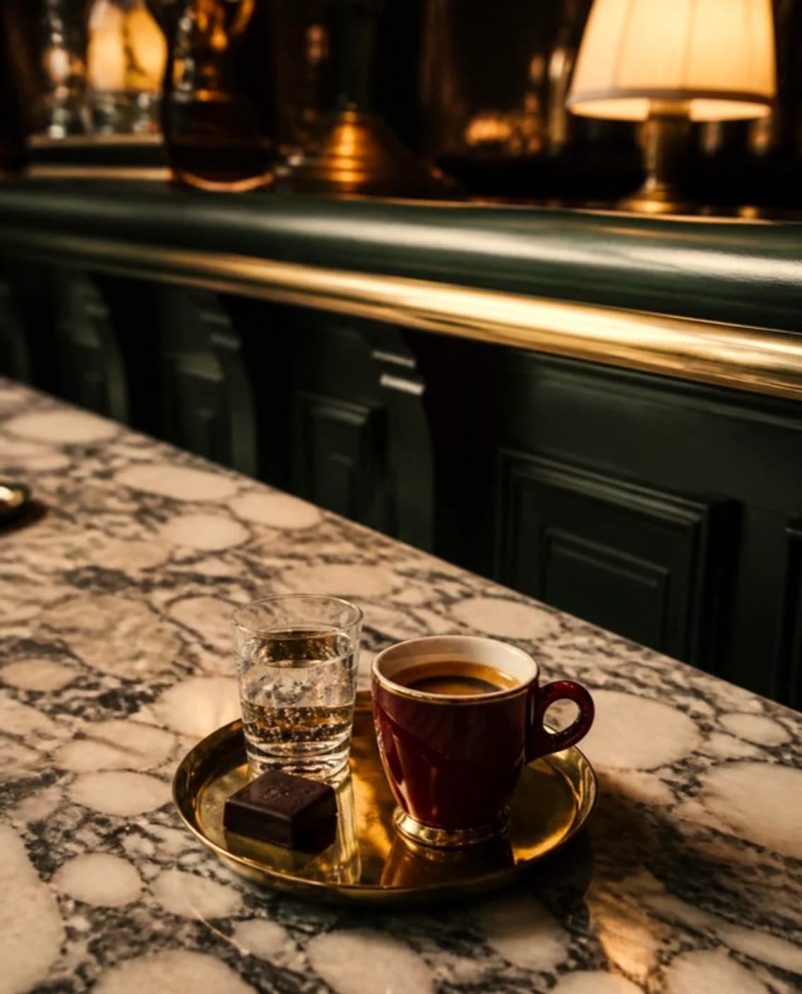 A small coffee in an oxblood cup on a brass tray, beside a glass of water and a square of chocolate